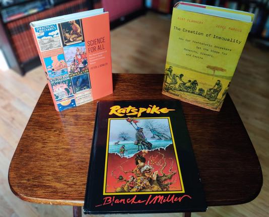A photo of three books standing on a small, brown, wooden table. On the left, standing up, Science for All, showing a collage of vintage science book covers. In the middle, lying down, Ratspike, showing two surrealistic drawings of human figures by Ian Miller and John Blanche. On the right, standing up, The Creation of Inequality, showing a yellow-tinted drawing of human figures, some naked, some wearing loincloths, in a savannah landscape. Most are kneeling in a large circle and have geometric figures painted on their chests, with some seemingly having some sort of bags or coverings on their heads and faces. Several other figures are standing behind the kneeling figures, bent over them and holding on to the coverings. The out-of-focus background shows black shelves full of books and part of the beech-coloured laminate floor.