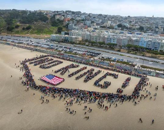10:25 in the morning on Ocean Beach, SF. [Scott Hess, San Francisco Bay Area Street Pbotography

Image shows an aerial photp of many people on the beah forming a large message which reads "No Kings" on top of a US flag (real flag held by people around it, not made up of people).