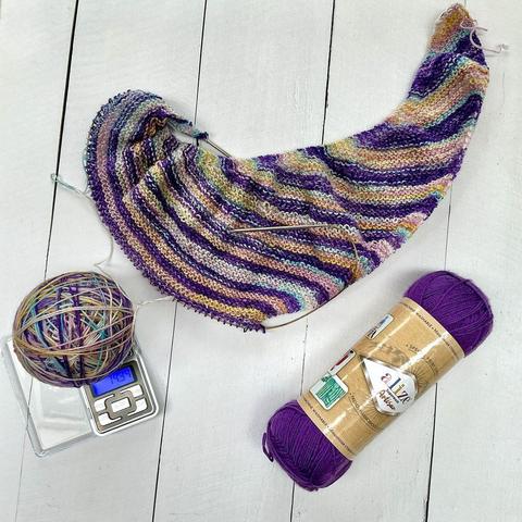An asymmetrical shawl project on a circular needle in garter stitch, it has stripes in various pastel colors. The ball of yarn lies on a small digital scale, there is also a skein of solid purple yarn.