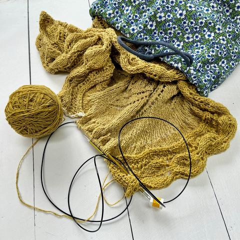A large light-green knitting project spilling out of a project bag, on a white plank background.