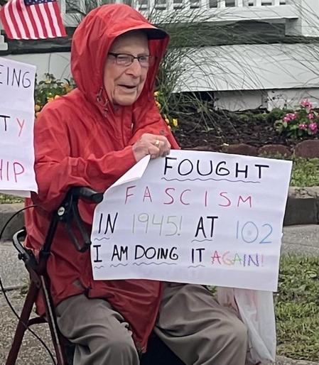 An elderly person wearing a red raincoat sits in a folding chair holding a sign that reads, "FOUGHT FASCISM IN 1945! AT 102 I AM DOING IT AGAIN!" A small American flag is visible in the background,