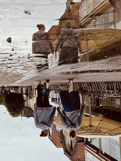A couple of two women walking on a cobble stone street, reflected in a puddle on the street. on the right side of the picture there are some chairs and tishes in front of a cafe, and yellow sun shades. The whole picture is turned upside down.