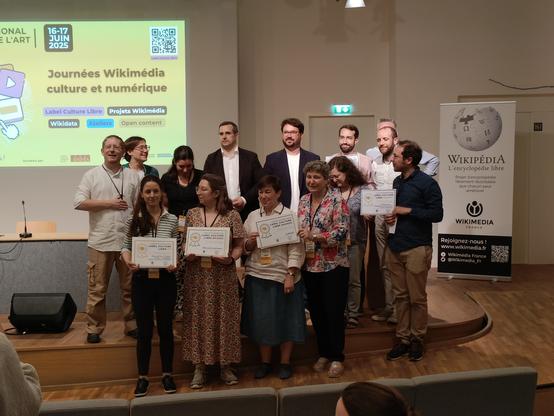 Photographie numérique couleur en format paysage.
Photographie de groupe du jury et des labellisés. Un écran à gauche et un kakémono Wikimedia France à droite.