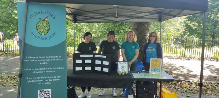 4 women standing behind a table in a market stall, a sign beside them read "Glasladies Beer Society " and links for more information.