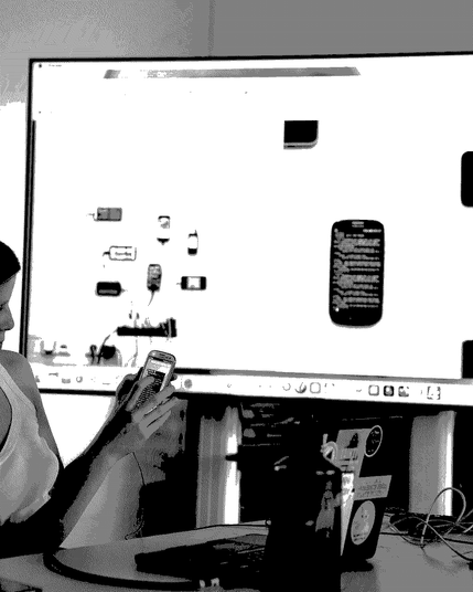 A person holding an older low-end smart phone. In the background there's a big screen showing similar phones plugged in and stuck to a wall, cryptically, aesthetically.