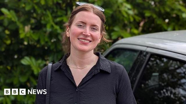 A woman smiles in a black dress next to a grey car