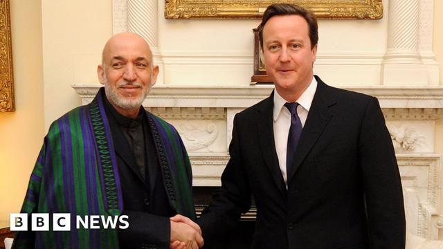 A meeting at Downing Street in 2011 between UK Prime Minister David Cameron, wearing a dark suit and tie, and Afghan President Hamid Karzai, who is wearing a dark suit and shirt and an embroidered chapan, an Afghan coat or cape, with purple and green stripes. They are shaking hands as they stand in front of a marble fireplace and paintings in gilded frames