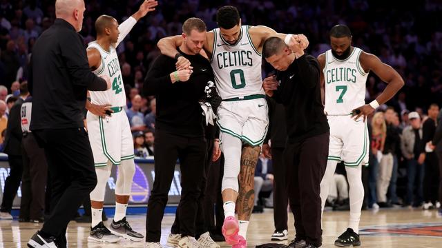 Jayson Tatum est soutenu par deux entraîneurs adjoints pour marcher, tandis que Jaylen Brown, la tête baissée, apparaît en arrière-plan.