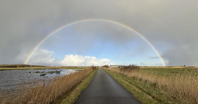 Im Zentrum verläuft ein gerader asphaltierter Weg, darüber ein Stück blauer Himmel, das von einem intensiven Regenbogen umrahmt wird. Außerhalb des Bogens ist der Himmel von grauen Wolken bedeckt.