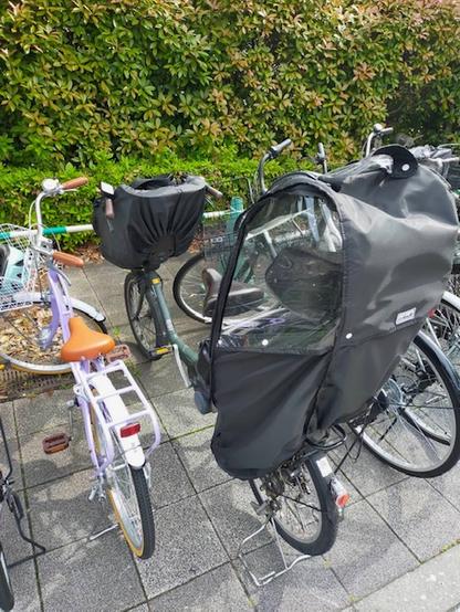 Photo is of a parked black "mamachari". It has a child seat in front and a child seat in back covered in rain-protecting plastic. To its left is a child's light purple bicycle with orange seat and handlebar grips. A green bush can be seen in the back.