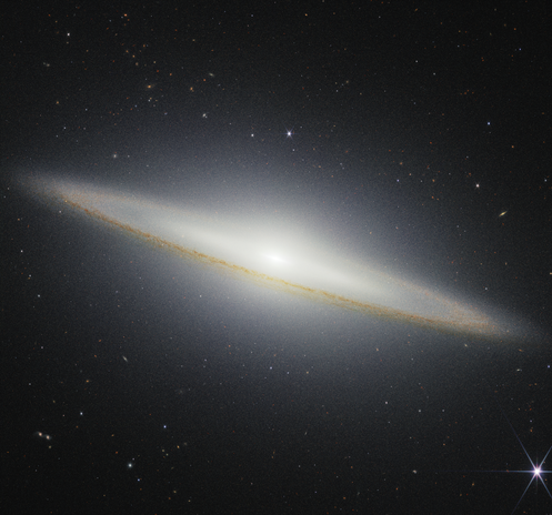 An observation of the Sombrero galaxy. The galaxy is a very oblong, brownish yellowish disk that extends from left to right at an angle (from about 10 o’clock to 5 o’clock). The galaxy’s center glows white and extends above and below the disk.