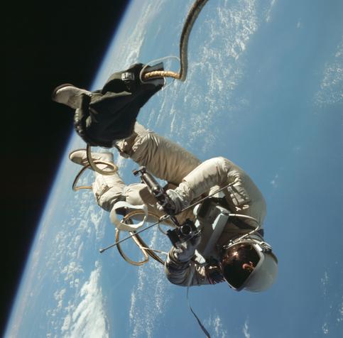 An astronaut in a spacesuit floats almost upside down outside of a spacecraft. Earth's blue water and white clouds are behind the astronaut.