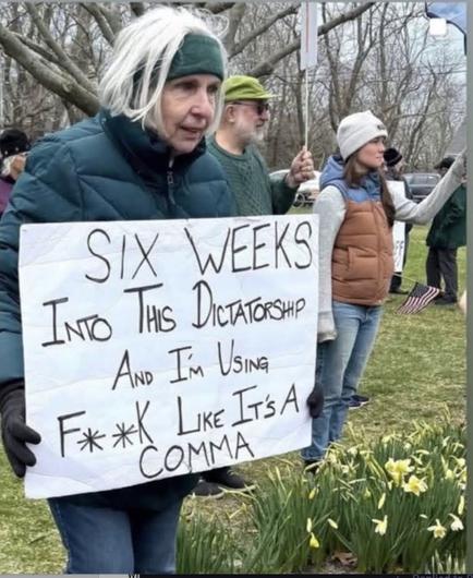 White-haired woman holds a sign reading SIC WEEKS INTO THIS DICTATORSHIP AND I'M USING F**K LIKE IT'S A COMMA