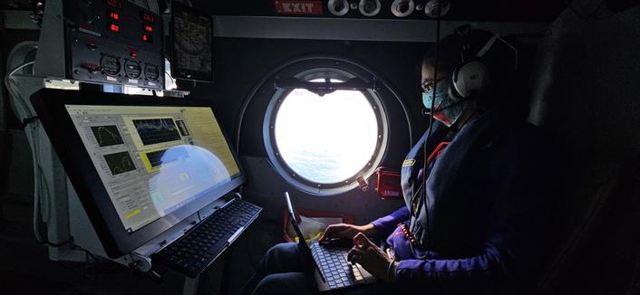 A woman in a blue facemask sits in an airplane, typing on a laptop with a large screen in front of her. The scene is lit by a round window in the middle of the frame. A woman in a blue facemask sits in an airplane, typing on a laptop with a large screen in front of her. The scene is lit by a round window in the middle of the frame.