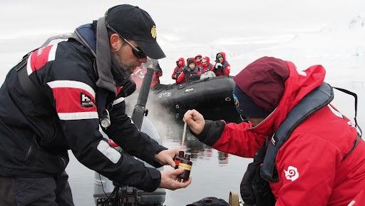 Scientists in cold-weather gear collect a water sample from a small boat in a polar region, with others observing from a nearby inflatable boat.