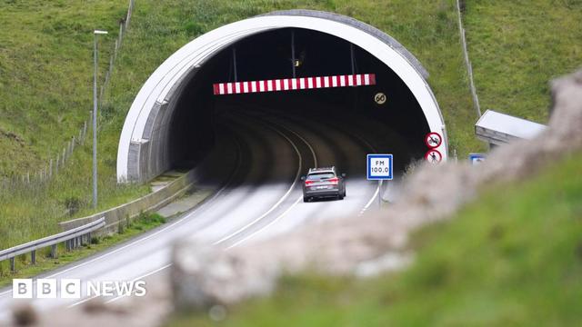 A grey car enters a tunnel, driving past red "no pedestrian" and "no cyclist" roadsigns. The tunnel disappears into a grassy hillside. A blue and white radio station information sign reads "FM 100.0".