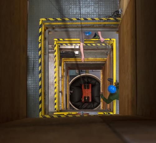 Looking down into the drop tower, two workers in hard hats stand on a platform halfway down. They watch a large red box suspended on a thick wire land into a black pillow-type material at the bottom of the test area.
