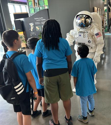 An astronaut mascot talks to a group of children wearing blue shirts. They are inside a building that highlights cultural information on Omaha, Nebraska.