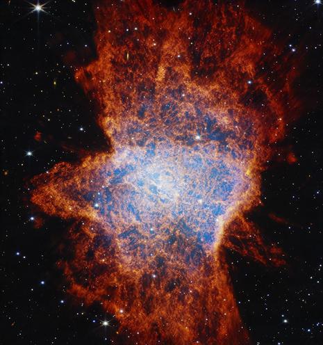 Colorful, mostly red glowing cloud with a distorted, asymmetrical shape that is illuminated from within by a bright central star. The asymmetrical shape resembles a large squished bug on the ground. In the center, a light blue glow appears over areas of dark pockets that look dark blue and are traced with orange material. It has a clumpy appearance. Shells of gas and dust appear as lobes stretching from roughly 11 to 5 o’clock, another from 1 to 7 o’clock, and possibly a third from 12 to 6 o’clock. The shells become a deeper red with distance from the center. These outflows push gas toward the equatorial plane, forming a disk that appears to span from 9 to 3 o’clock. The background of space is black and speckled with tiny bright stars and distant galaxies.
