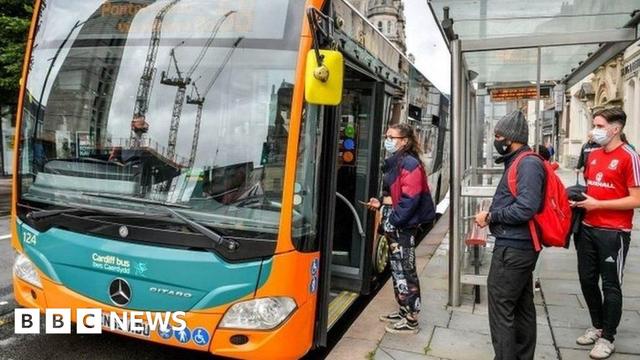 Three people wearing masks queuing to get on to a Cardiff Bus, parked up at a bus stop. The bus is a single decker and has the company's logo on the front.