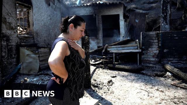 A lady with black hair wearing a black vest walks with her head down next to a blackened and charred burnt tavern
