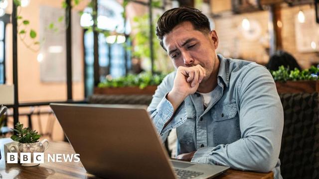A young man with short dark hair and wearing a denim shirt appears distracted sitting in front of a laptop, which is on a wooden table, in a large open space. In the blurred background you can make out ceiling lamps, climbing plants and other wooden tables.