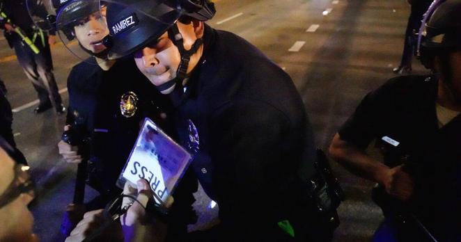 An LAPD officer swings a baton at photojournalist Nicholas Stern as he displays his press credential during an immigration protest in Downtown L.A. on Aug. 8, 2025.