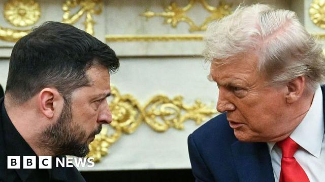 Zelensky and Trump in conversation in Oval Office. Trump is wearing a blue suit and red tie, Zelensky a black suit jacket. They both have a serious expression.