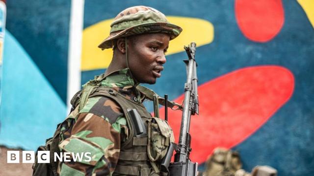 A soldier wearing army fatigues and carrying a gun stands in front of a colourfully-painted wall.