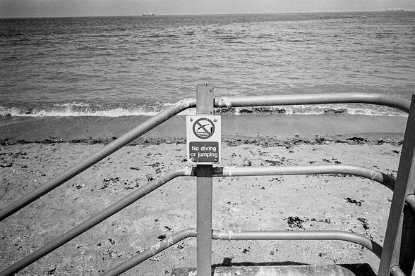 a view from a walkway to a beach, a sign depicting a person diving into the water reads 'no diving or jumping'