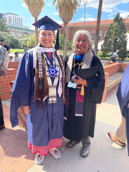 Two women are standing on the mall at UArizona in graduation regalia.  One is also wearing Northern Cheyenne regalia, with incredibly beautiful beadwork.  The other has a beautiful red and blue sash.  They are smiling and beaming with pride, which is well deserved.  