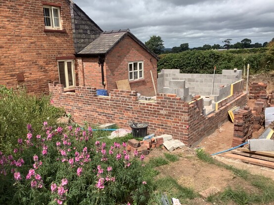 Looking at the corner of a red brick house, an extra room is being built on to it, the walls half constructed up to window level