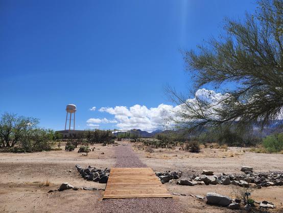 a desert expanse with a wooden bridge over a little wash, some palo verde trees, and a water tower in the background.  The sky is bright blue but there are puffy white clouds rolling in that might drop a little rain this evening.