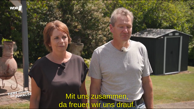 Zwei Personen stehen im Freien vor einem Gartenhaus. Untertitel: 'Mit uns zusammen, da freuen wir uns drauf.'