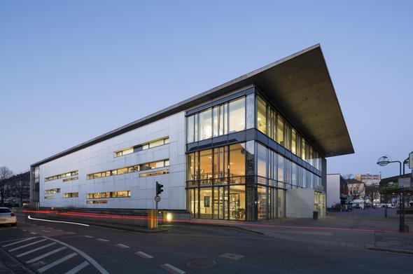 Außenansicht der Mediothek Krefeld in der Abenddämmerung. Das moderne Gebäude besitzt eine helle Fassade, große Glasfronten und ein weit ausladendes Dach. Durch die beleuchteten Fenster sind die Innenräume zu erkennen.