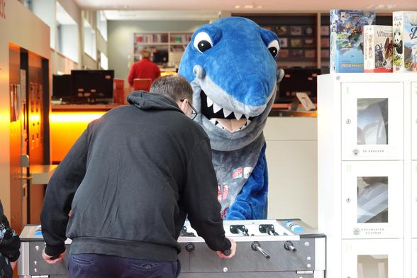 Ein Mann spielt an einem Tischkicker in der Mediothek Krefeld. Ihm gegenüber steht eine Person in einem großen, blau-grauen Hai-Kostüm mit weit geöffnetem Maul und weißen Zähnen. Im Hintergrund sind Computerarbeitsplätze und Regale mit Gesellschaftsspielen zu sehen.