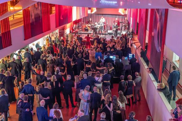 Blick von oben auf eine gut besuchte Veranstaltung in der Mediothek Krefeld. Zahlreiche Menschen stehen vor einer Bühne, auf der eine Band auftritt. Viele Gäste haben sich untergehakt und tanzen gemeinsam. Rote Vorhänge und farbige Beleuchtung prägen die Atmosphäre.