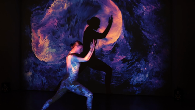 Three female performers in white dance outfits stand in a projection beam that paints colorful fractal clouds of bright orange and dark blue light onto them and the back wall. The colorful clouds may remind of natural phenomena like flames, flowing lava, smoke or corals. The software of the interactive artwork that is used to create the projected clouds allows the clouds to react to the movement of their shadows, so both sound and visuals of the projection seem to magically adapt to the dancer's presence and flow along with their movements.