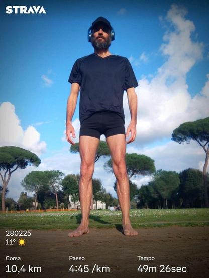 Un uomo barbuto con cuffie e berretto indossa una maglietta e pantaloncini neri, a piedi nudi su un sentiero sterrato in un parco alberato. In sovraimpressione compaiono dati di Strava relativi a una corsa: 10,4 km, passo di 4:45/km, tempo di 49 minuti e 26 secondi.