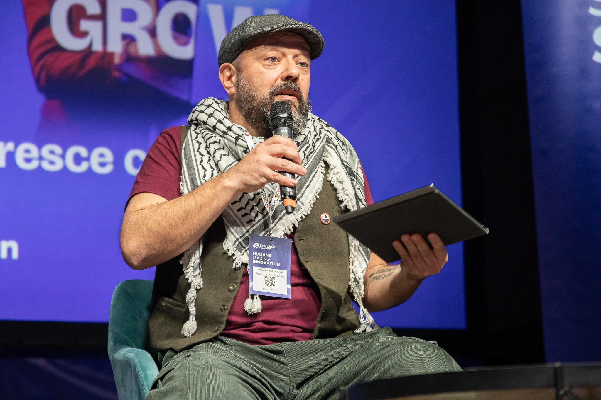 Morloi sul palco di SMStrategies, impegnato a spiegare il fediverso. Porta una coppola, una Kefiah e la spilletta di BadTaste/ZTL. 