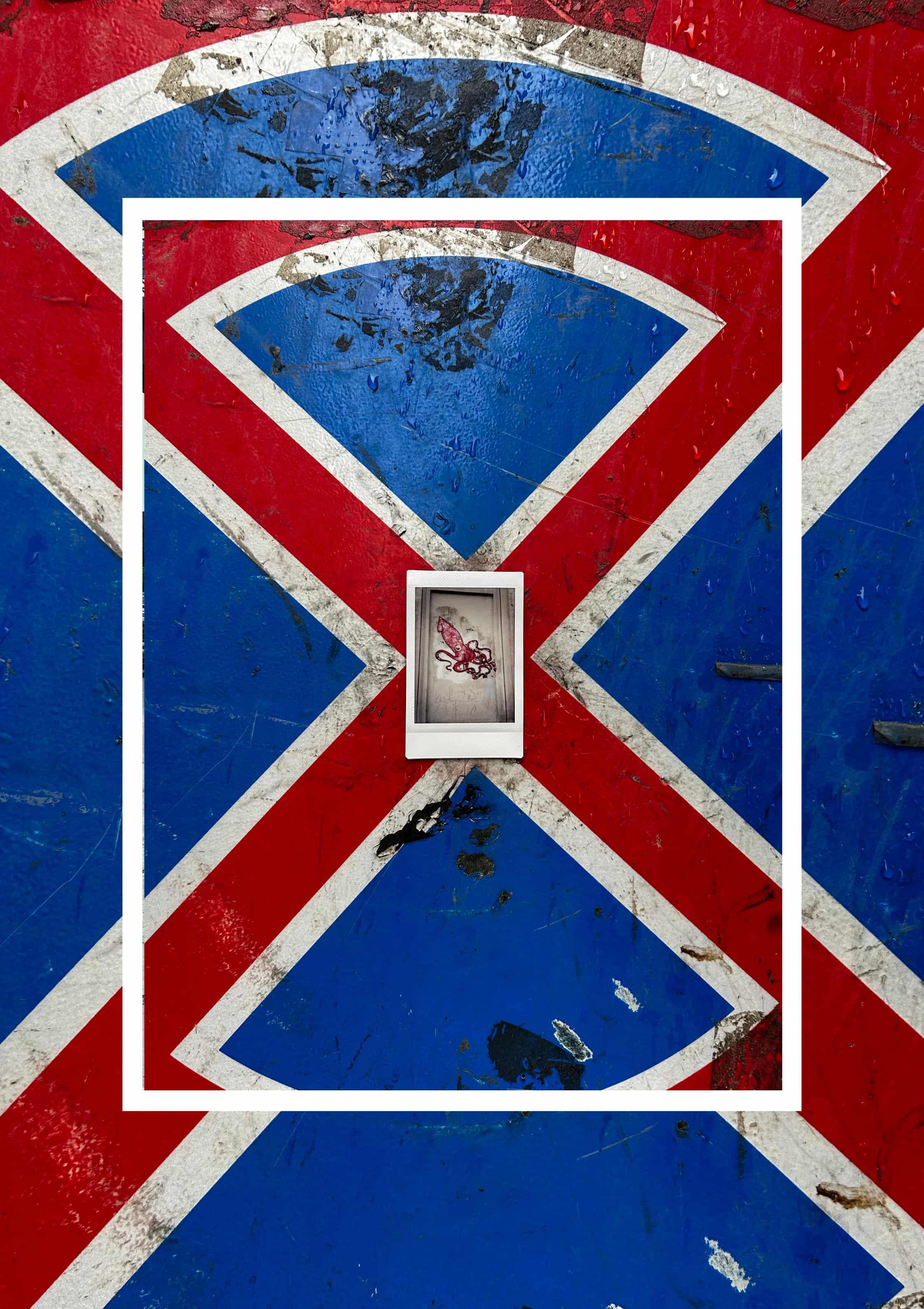 Small white instant photo centered on a scratched red-and-blue painted surface with white diagonal striping; a thick white rectangular border frames a smaller inset view of the same patterned background, creating a nested effect.