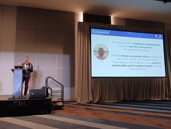 Photo of Maciej on stage, next to a big slide that says "Sustainability" with a quote by Dims.