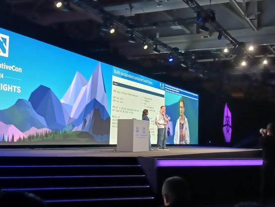 Kubecon keynote stage photo.  Colin Walters, Preethi Thomas, and Dan Walsh on stage.  Behind them is a demo of Bootc updating system images.