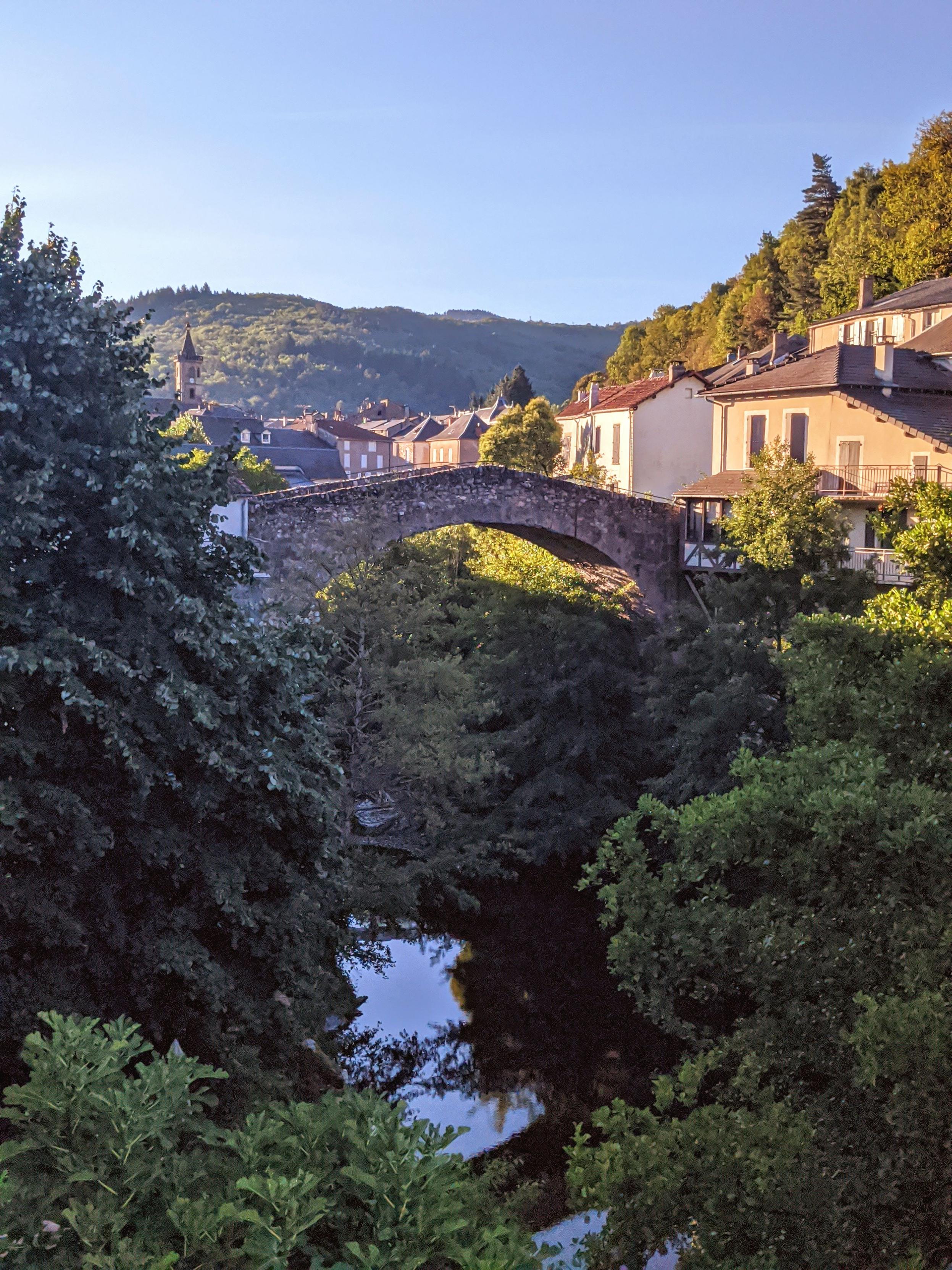 Un vieux pont de pierre médiéval  au-dessus d'une petite rivière. On est dans un village de l'Aveyron, entouré de collines et de forêts.