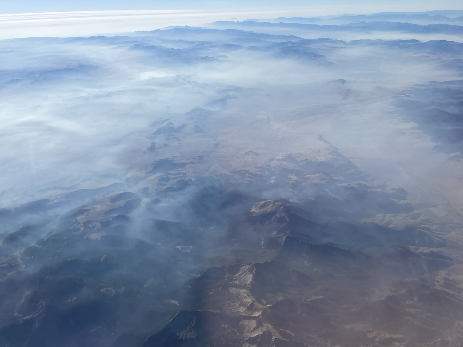Aerial view of a mountain range shrouded in clouds and fog, showcasing layered hills under a soft blue sky.