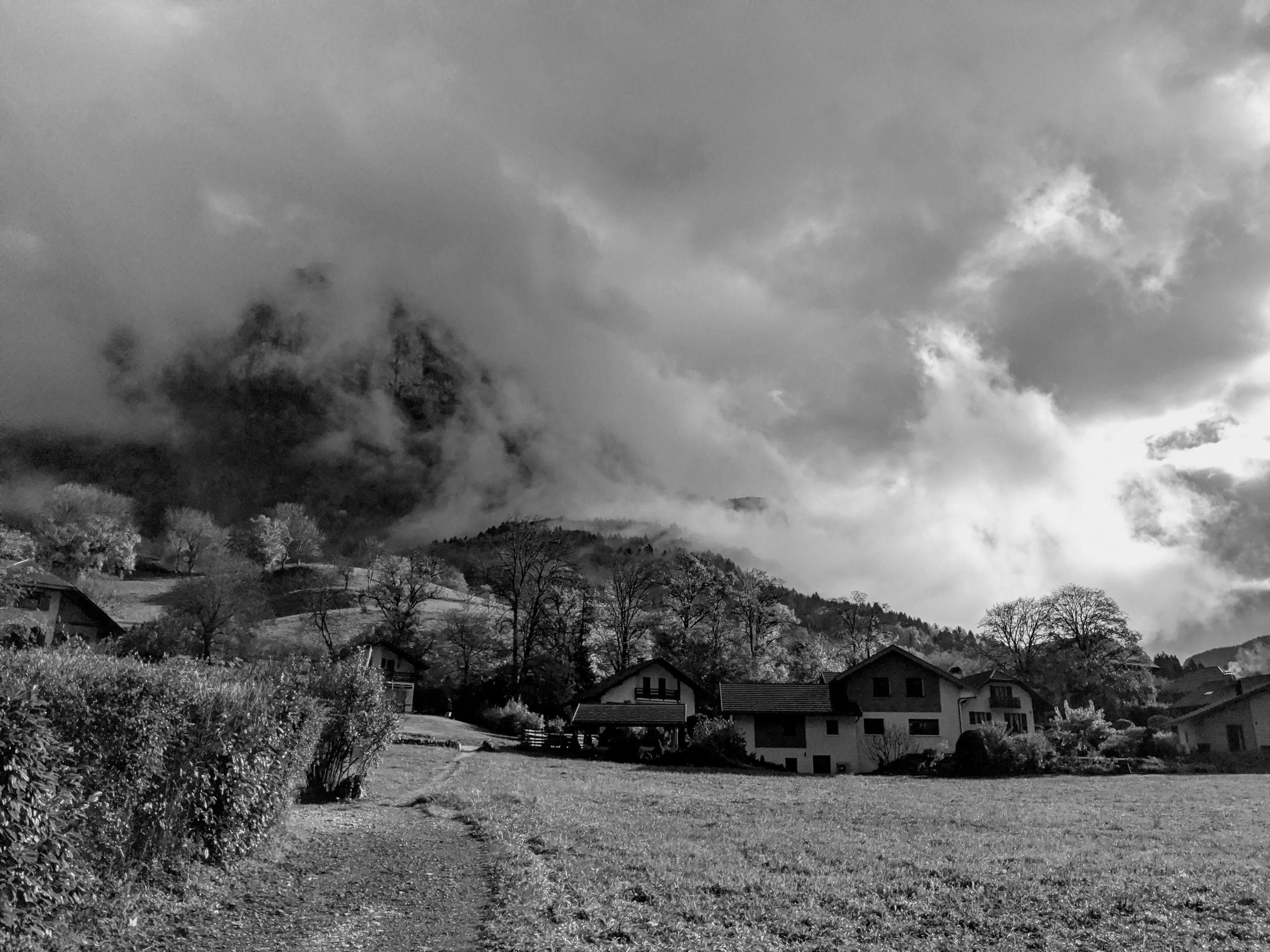 Un paysage montagnard en noir et blanc. La montagne disparaît dans des nuages qui s'effilochent le long de la pente. Au premier plan, un champ d'herbe et quelques chalets.