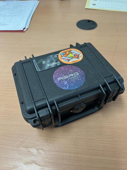 A small black protective case with a handle sits flat on a table. There are three stickers on the box, one labelled ASRG for the Astromaterials Science Research Group at JAXA, an orange Hayabusa2 mission logo sticker, and another Hayabusa2 sticker showing an artist impression of the spacecraft.