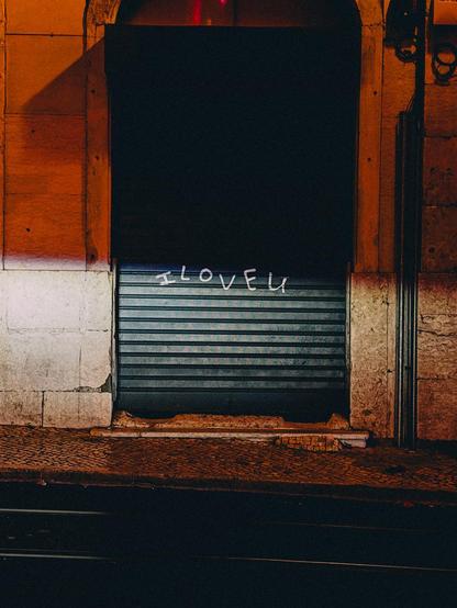 Closed storefront with dark metal shutters and graffiti text reading “I LOVE U” on a dimly lit street at night.