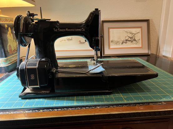 A Singer Featherweight on a table. Behind and to the left is a small lamp and behind are two framed prints of birds.