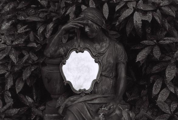 Black and white film photo of a statue of a woman in a thinking pose, an antique brass mirror placed upon her lap, a bush with speckled leaves around her.